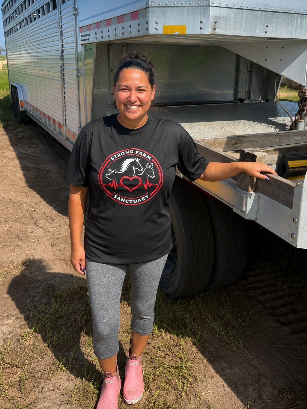 Supporter wearing a black Lock Pon Stop Strong Farm Sanctuary T-shirt featuring a heart, horse, cow, and rabbit logo while standing outdoors beside a livestock trailer at the sanctuary. This animal rescue fundraiser shirt helps support the care and protection of rescued farm animals.