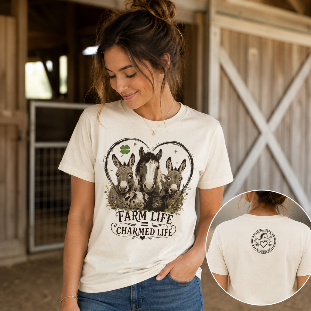 Lifestyle photo of a woman wearing the white Farm Life Charmed Life Strong Farm Sanctuary support shirt in a rustic barn. The front features a vintage-style rescued animal graphic, while an inset image shows the Strong Farm Sanctuary logo printed below the back collar.